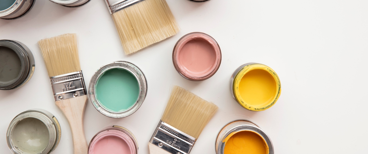 A collection of open paint pots of bright colours with several paintbrushes