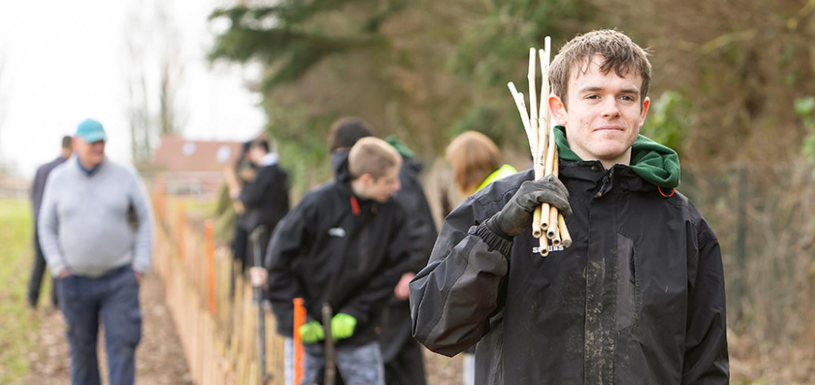 In the forefront of an image a teenage boy is carrying bamboo sticks over his shoulder whilst there are other people in the background in a field walking behind him and planting trees