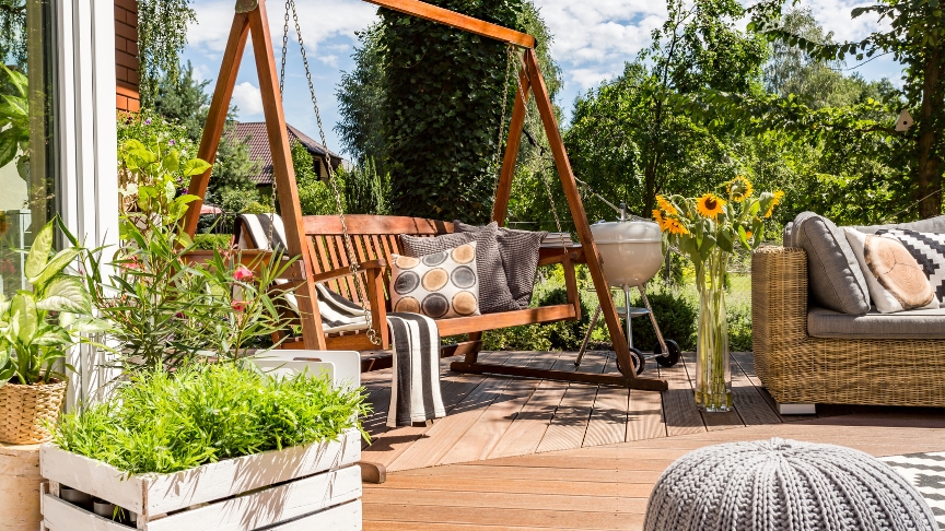 A wooden decking area with a swing seat, sunflowers, a barbecue and more plants