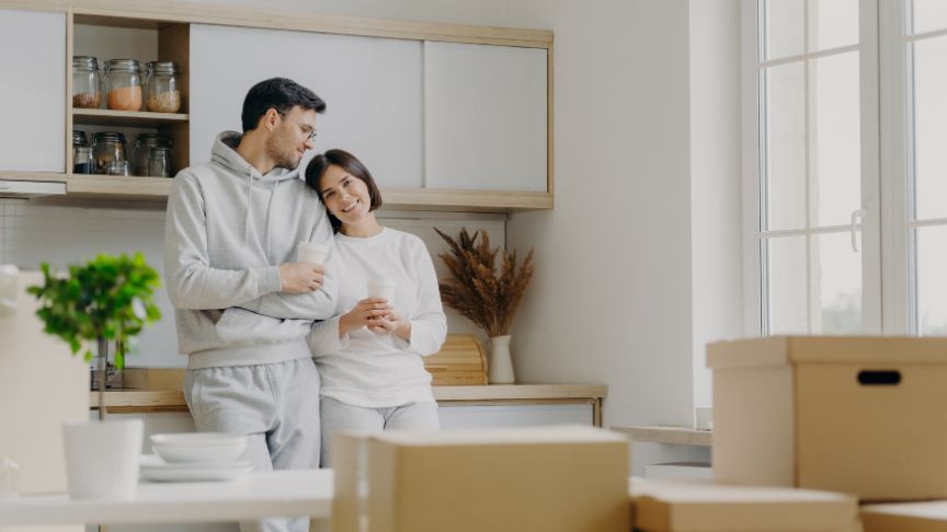 A female stood leaning on a male against a kitchen worktop looking at moving boxes.