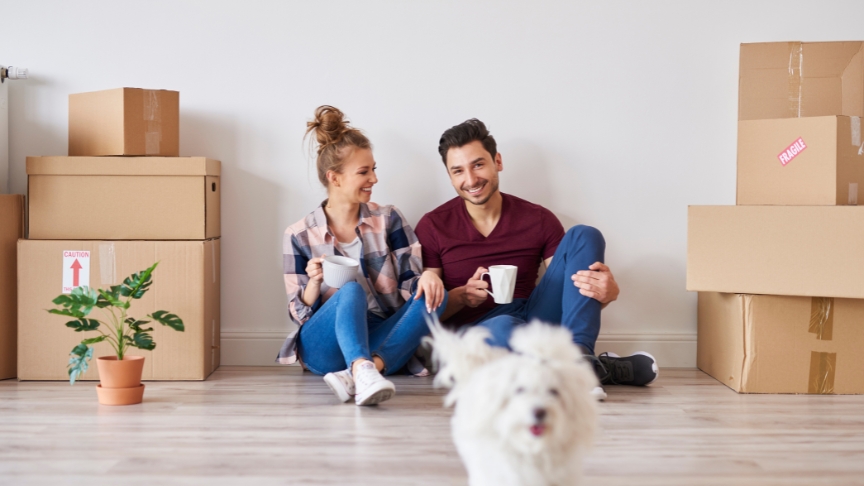 A couple sat on the floor of their new home with boxes beside them. They have a mug in their hand and look happy. Their small white dog is running towards the camera in front of them