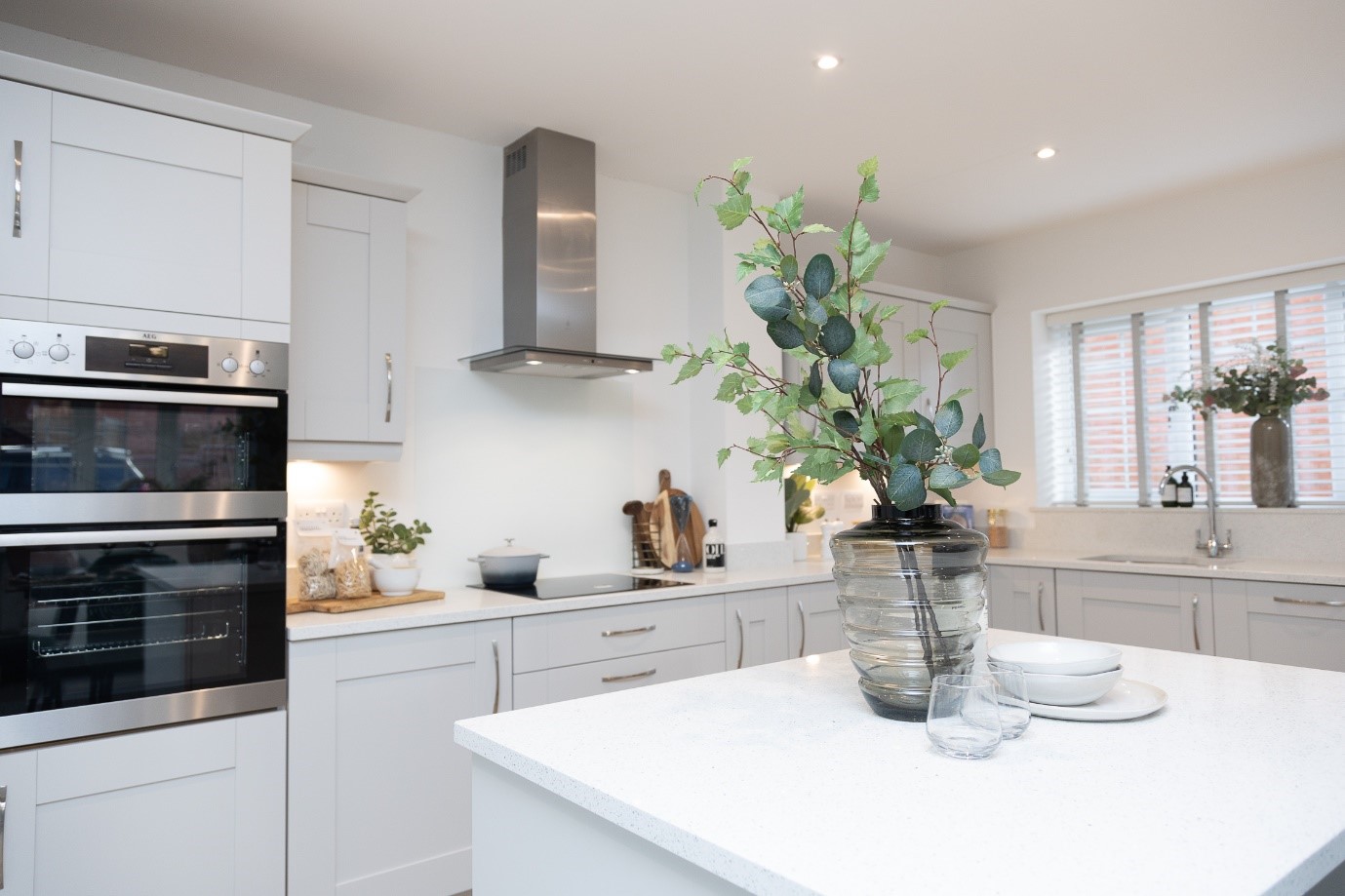 Interior of a clean and tidy white kitchen with cream cabinets. In the foreground is a kitchen island on which is a vase with foliage in and some crockery next to it