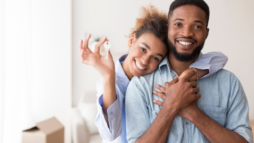A male and female couple smiling at the camera, with the female holding up a key. She has her arm over the male in a sweet embrace