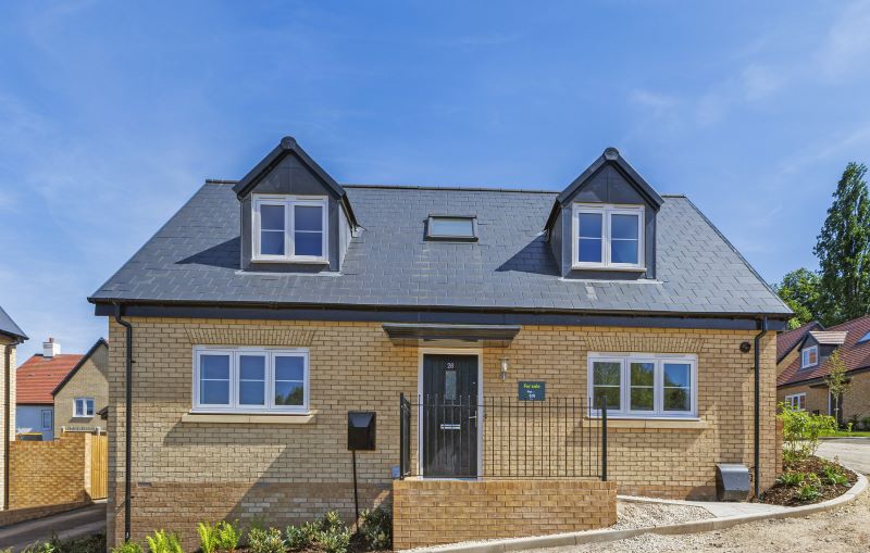 Modern two-story detached home with light brick exterior, dark slate roof, dormer windows, landscaped front garden, and central black front door at Coppice View..