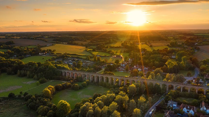 Aerial view of a countryside viaduct at sunset, with green fields, trees, and scattered houses.