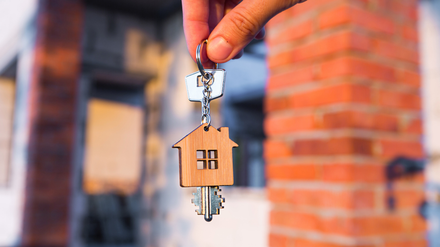 A house key with a home shaped wooden key ring attached are being held by two fingers. In the blurred background you can see a red bricked home.