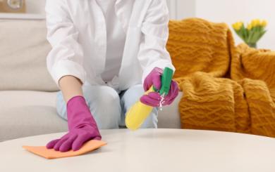 A person with yellow marigold gloves on holding cleaning spray and a sponge cleaning a wooden coffee table