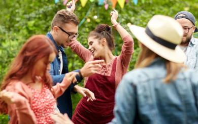 A group of men and women dancing in a garden decorated with multicoloured bunting.