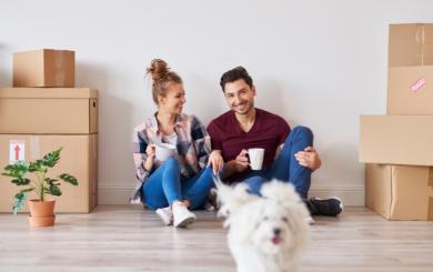 A couple sat on the floor of their new home with boxes beside them. They have a mug in their hand and look happy. Their small white dog is running towards the camera in front of them