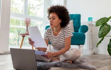 Stock image of a young woman sitting on a rug on a floor holding several pieces of paper and looking at a laptop