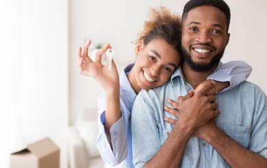 A male and female couple smiling at the camera, with the female holding up a key. She has her arm over the male in a sweet embrace
