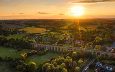 Aerial view of a countryside viaduct at sunset, with green fields, trees, and scattered houses.