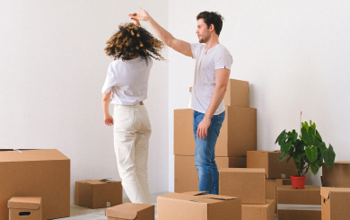 Two people in a room full of cardboard boxes, with one person playfully spinning the other by the hand. A green plant in a red pot is visible among the boxes.