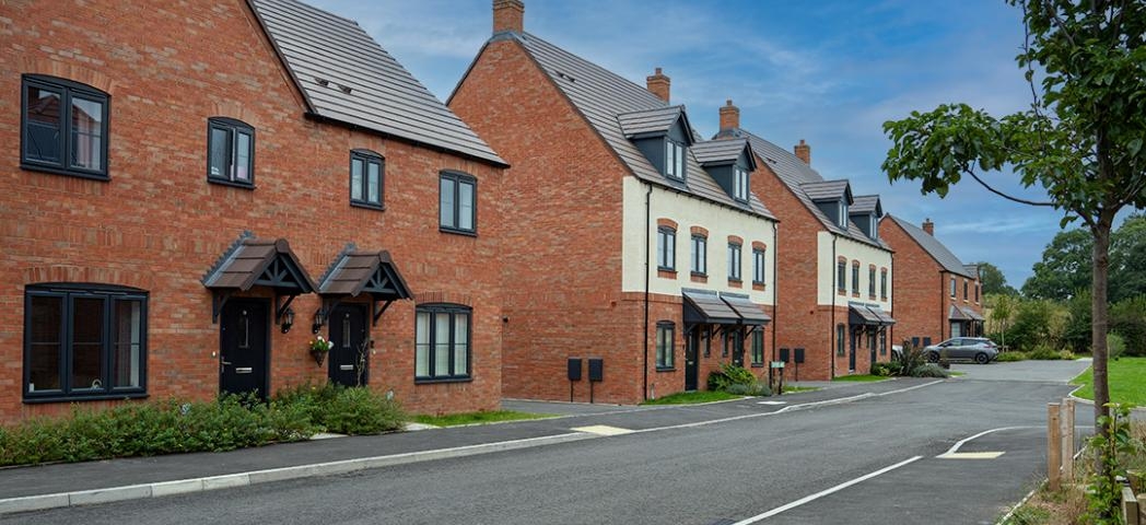 A row of modern, two-story red-brick townhouses with pitched roofs and black-framed windows, some sections featuring white plaster. Each house has a small front garden with shrubs, and they are aligned along a clean, paved street.