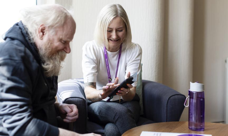 A woman with short blonde hair and a white shirt is sitting next to a man with a long beard, both smiling as they look at a smartphone together. They are seated on a couch in a well-lit room, with a water bottle and a document on a table in front of them. The atmosphere appears friendly and collaborative.
