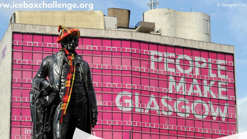 A large multistorey building in Glasgow with the windows covered pink with large writing that reads people make Glasgow. A statue of a man wearing a hat and scarf stands in the forefront