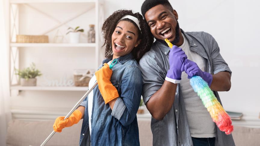 A young woman and man leaning on each other smiling and holding a duster and a brush