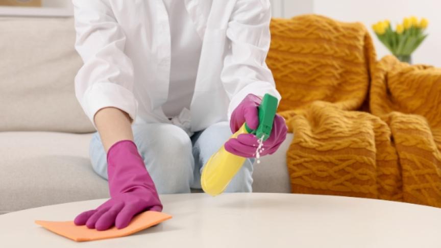 A person with yellow marigold gloves on holding cleaning spray and a sponge cleaning a wooden coffee table
