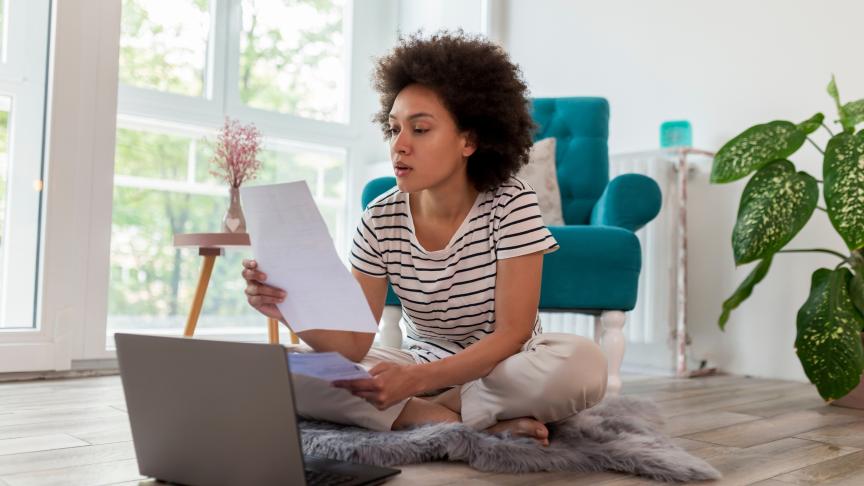 Stock image of a young woman sitting on a rug on a floor holding several pieces of paper and looking at a laptop