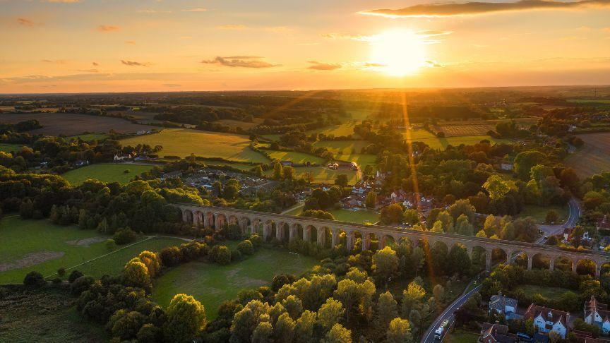 Aerial view of a countryside viaduct at sunset, with green fields, trees, and scattered houses.