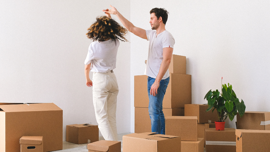 Two people in a room full of cardboard boxes, with one person playfully spinning the other by the hand. A green plant in a red pot is visible among the boxes.