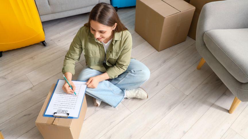 Woman sat on the floor and ticking off items from her moving day checklist while leaning on a closed box of possessions