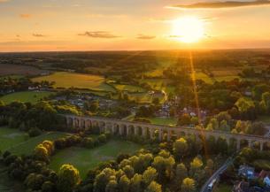 Aerial view of a countryside viaduct at sunset, with green fields, trees, and scattered houses.