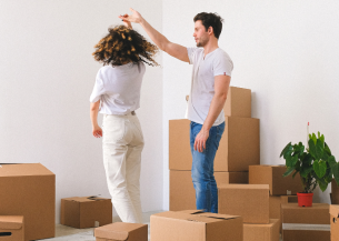 Two people in a room full of cardboard boxes, with one person playfully spinning the other by the hand. A green plant in a red pot is visible among the boxes.