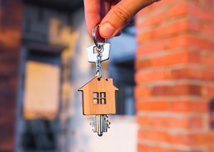 A house key with a home shaped wooden key ring attached are being held by two fingers. In the blurred background you can see a red bricked home.